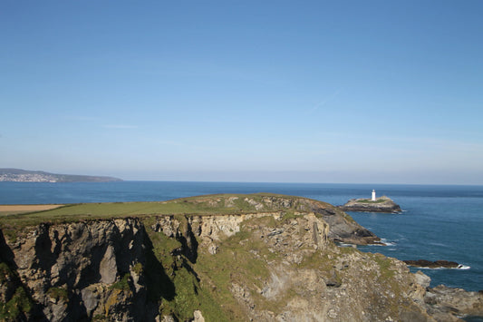 Rocky coastal cliffs with green grass and a white lighthouse on a small island in the ocean under blue sky