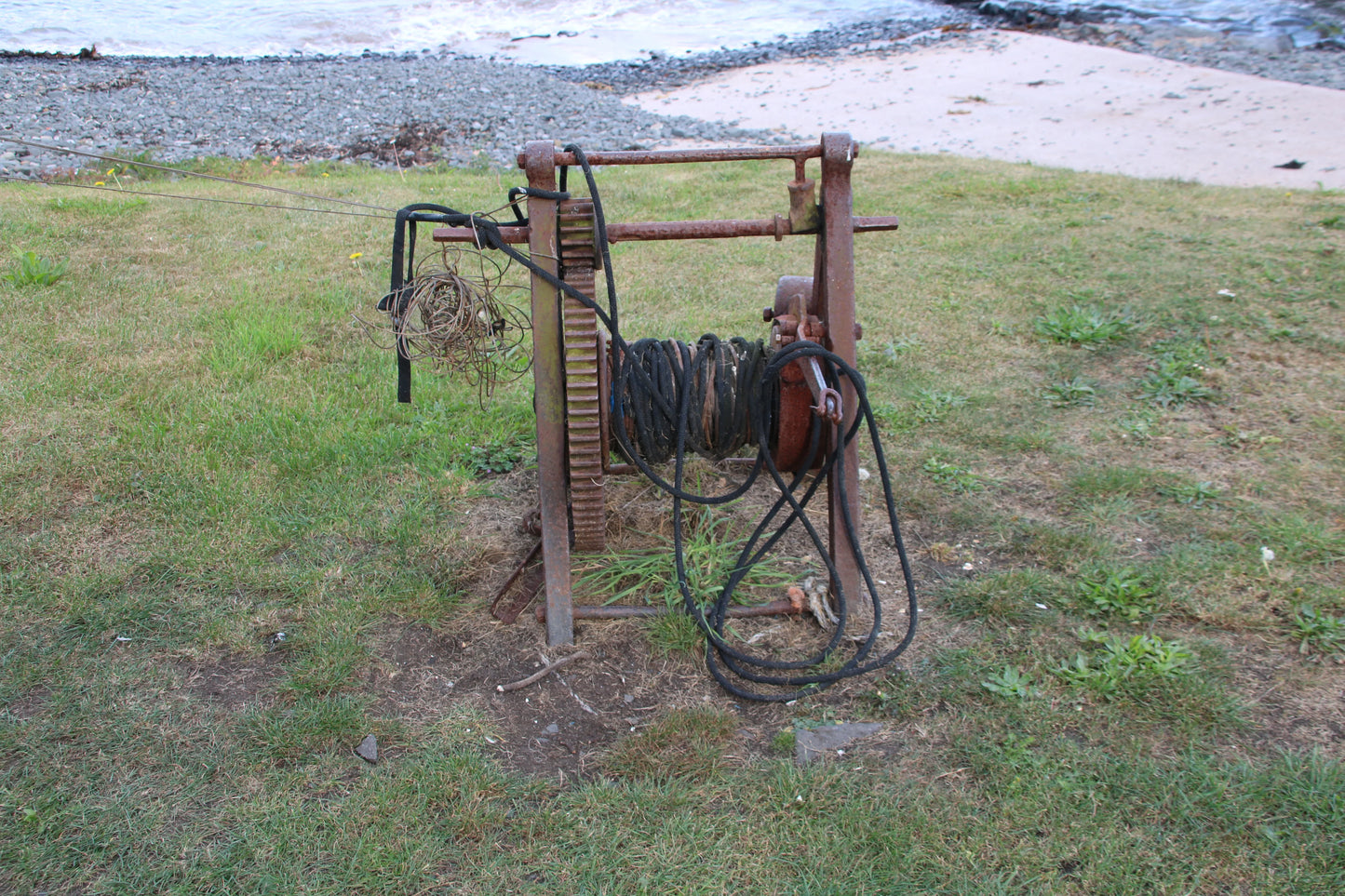 Rusty hand winch with coiled ropes on green grass by rocky shoreline