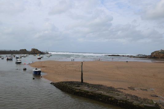 Coastal harbor with moored boats, sandy beach, rocky breakwater, and cloudy sky