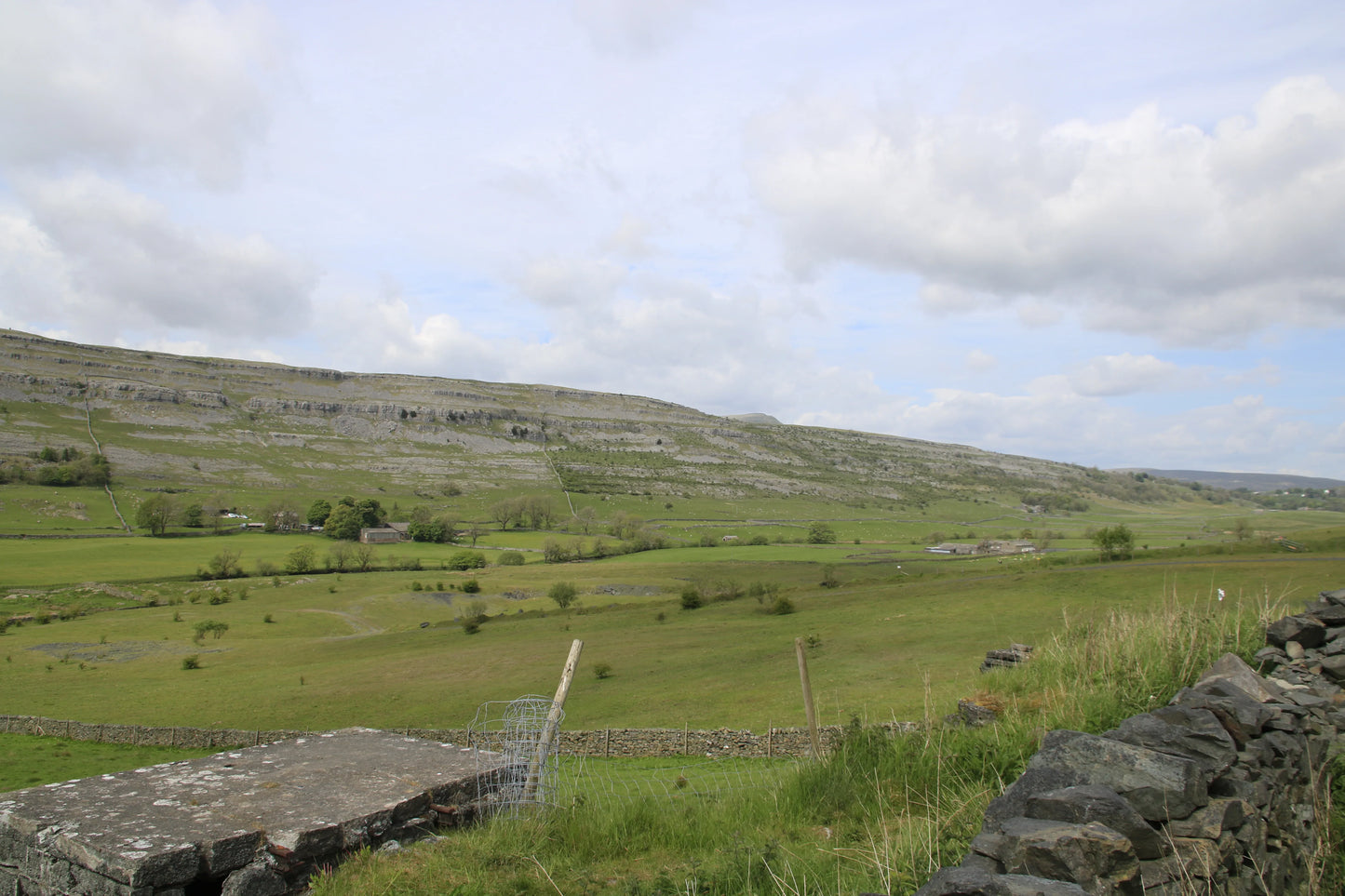 British rural landscape with green fields, drystone walls, scattered farm buildings, and a limestone hill under a cloudy sky
