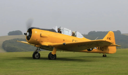 Yellow vintage military trainer airplane with two pilots taxiing on grassy airfield