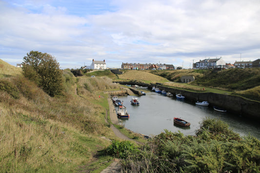 Small boats moored in a narrow harbor surrounded by grassy hills and houses under a partly cloudy sky
