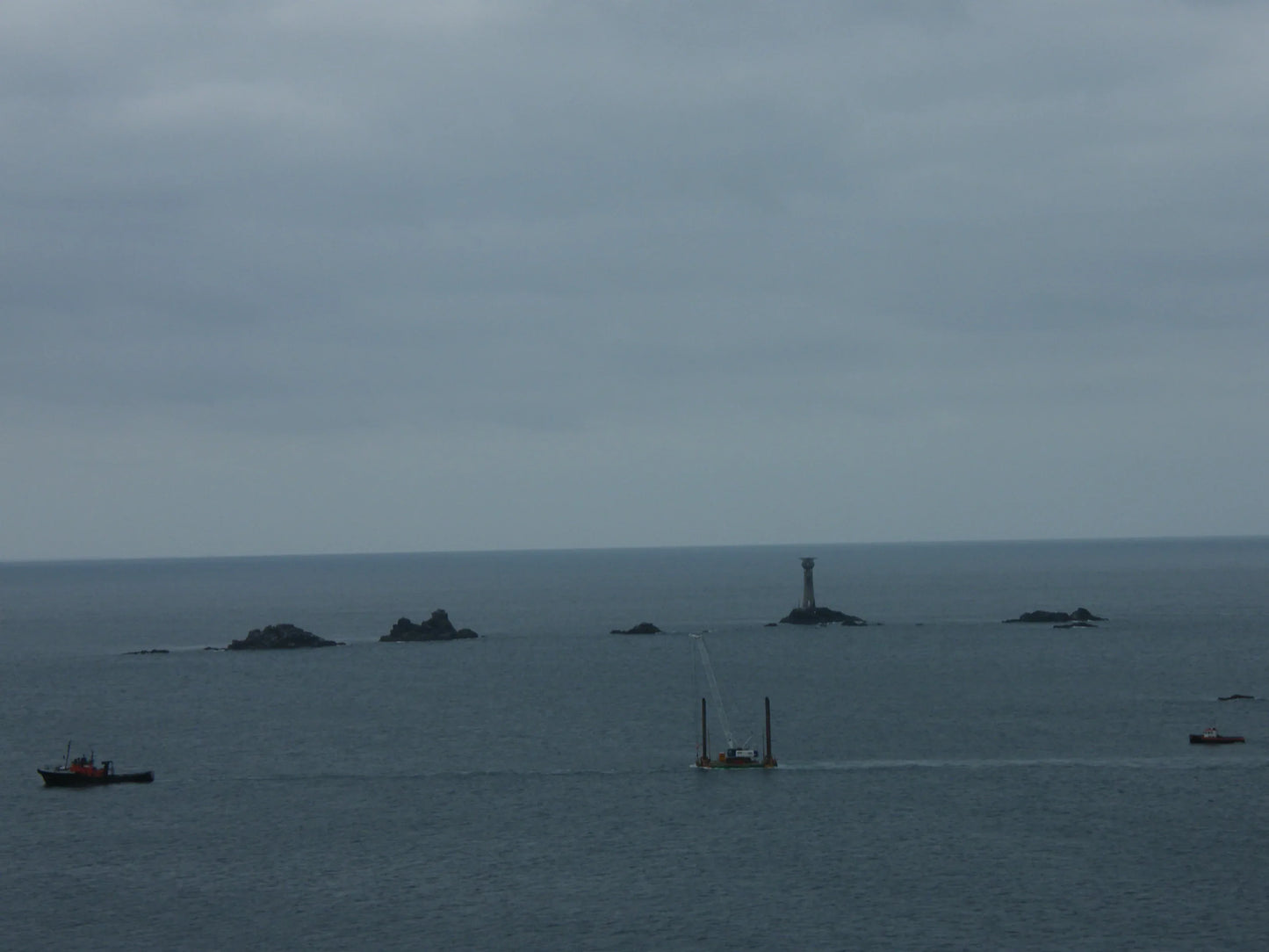 Overcast ocean scene with rocky islets and a tall lighthouse tower in the distance, vessels nearby