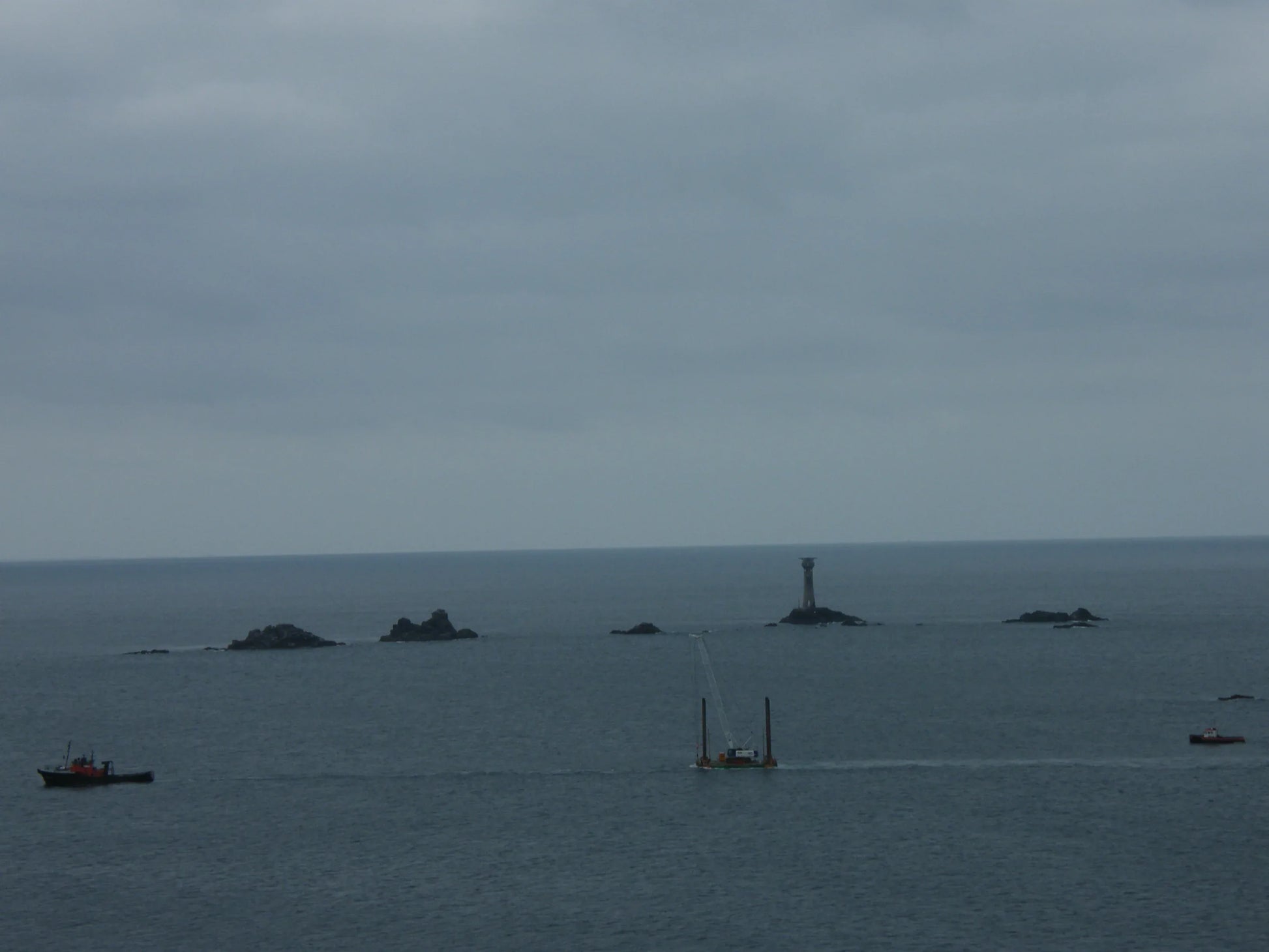 Overcast ocean scene with rocky islets and a tall lighthouse tower in the distance, vessels nearby