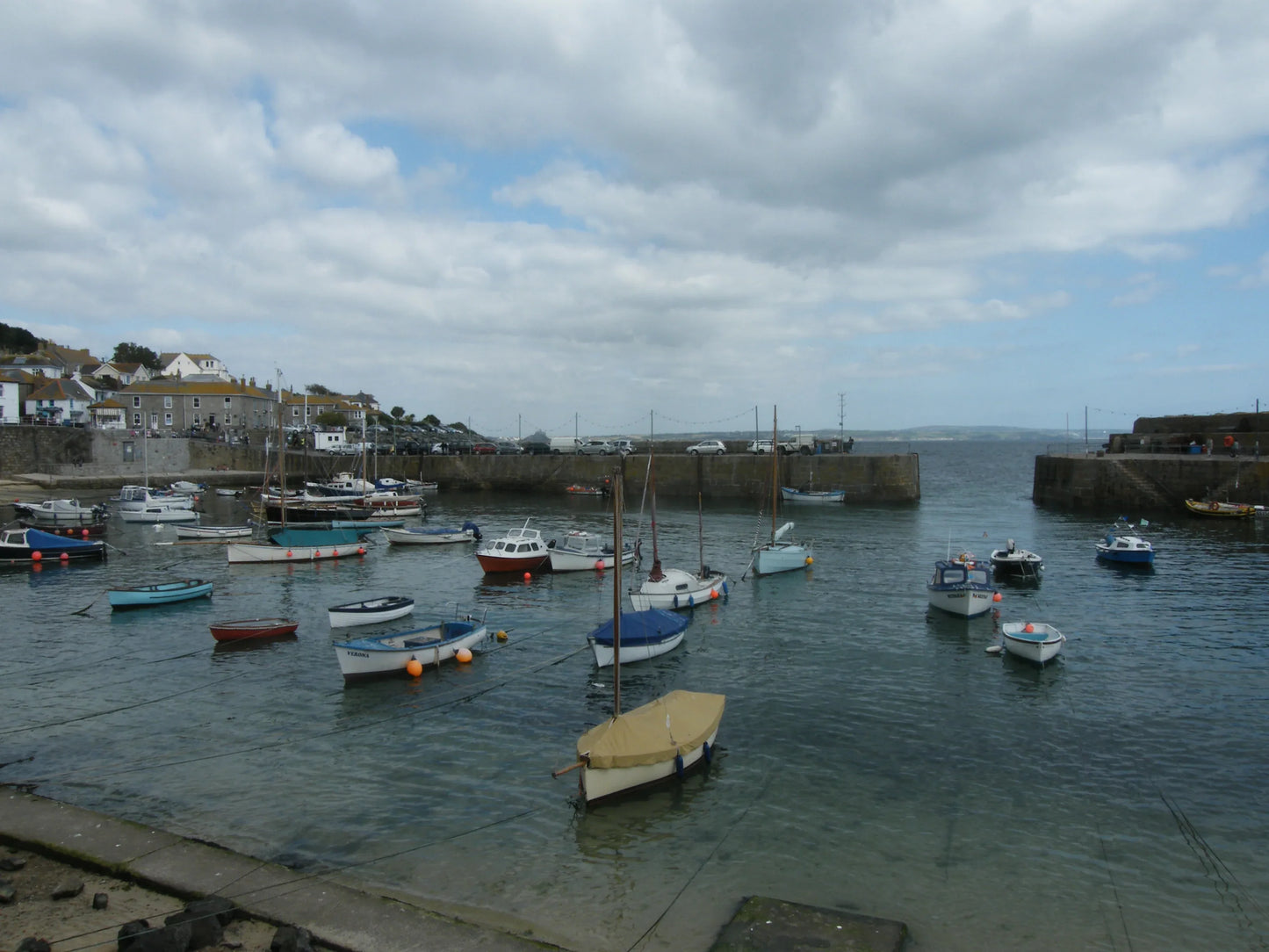Small sailboats and fishing boats moored in a calm harbor with stone piers and cloudy sky