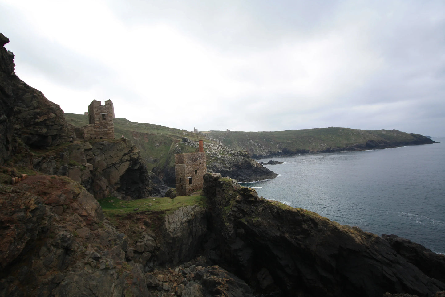 Historic stone mining engine houses on rocky cliffs overlooking calm sea at Cornwall coast under cloudy sky