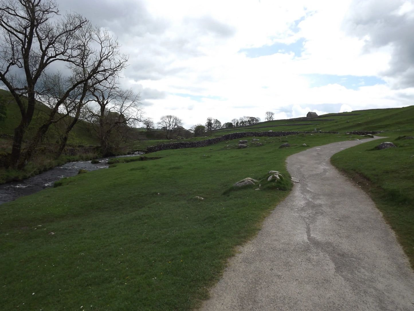 Curved country path beside a stream with leafless trees and green fields under cloudy sky