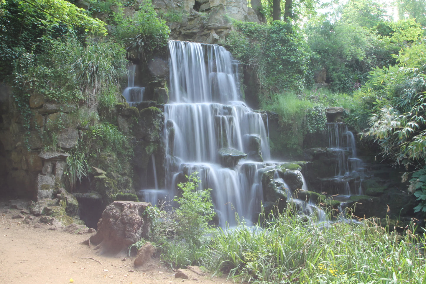 Multi-tiered waterfall cascading over mossy rocks surrounded by lush green plants in a forest setting