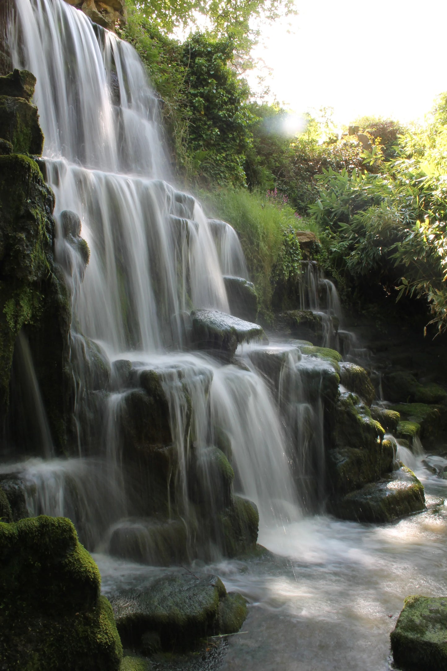 Multi-tiered waterfall cascading over moss-covered rocks surrounded by lush green foliage in sunlight