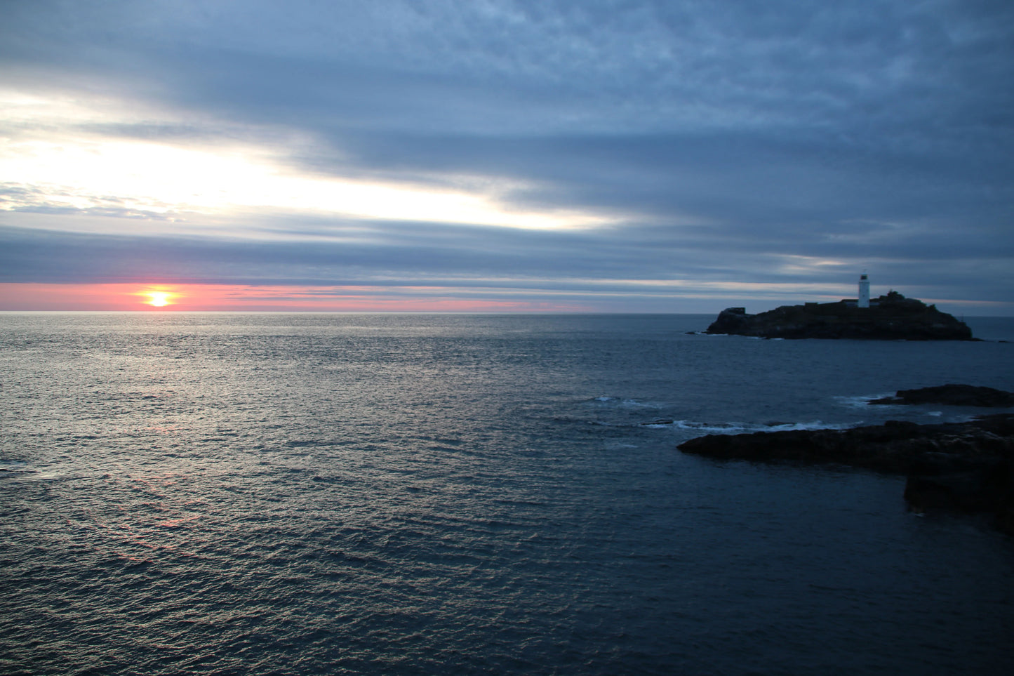 Sunset over calm ocean with lighthouse on rocky island under cloudy sky