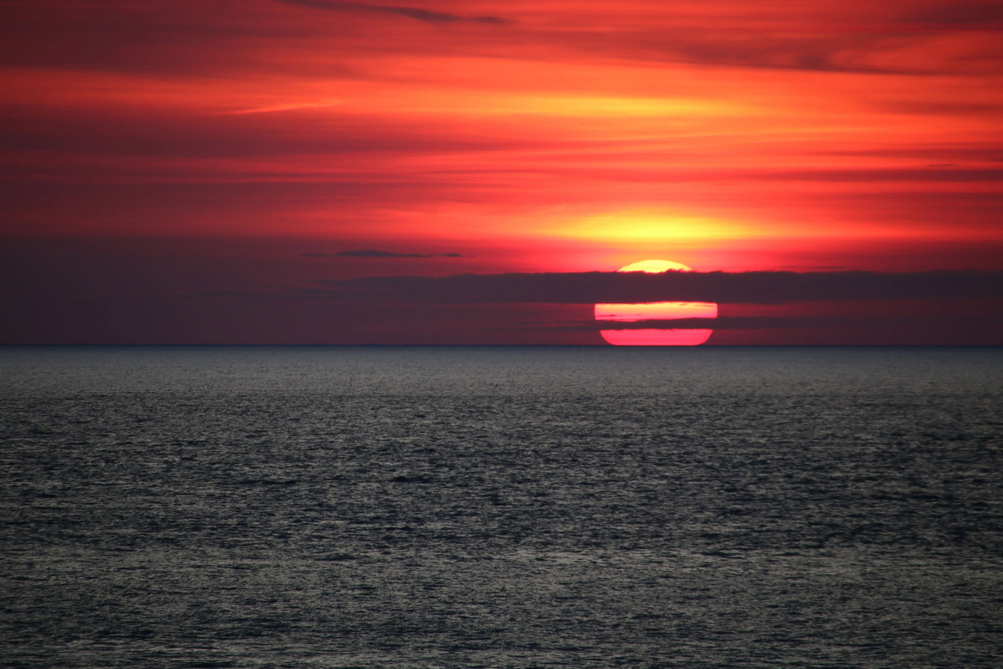 Sunset over calm ocean with red and orange sky and sun partially covered by clouds