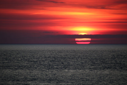 Sunset over calm ocean with red and orange sky and sun partially covered by clouds