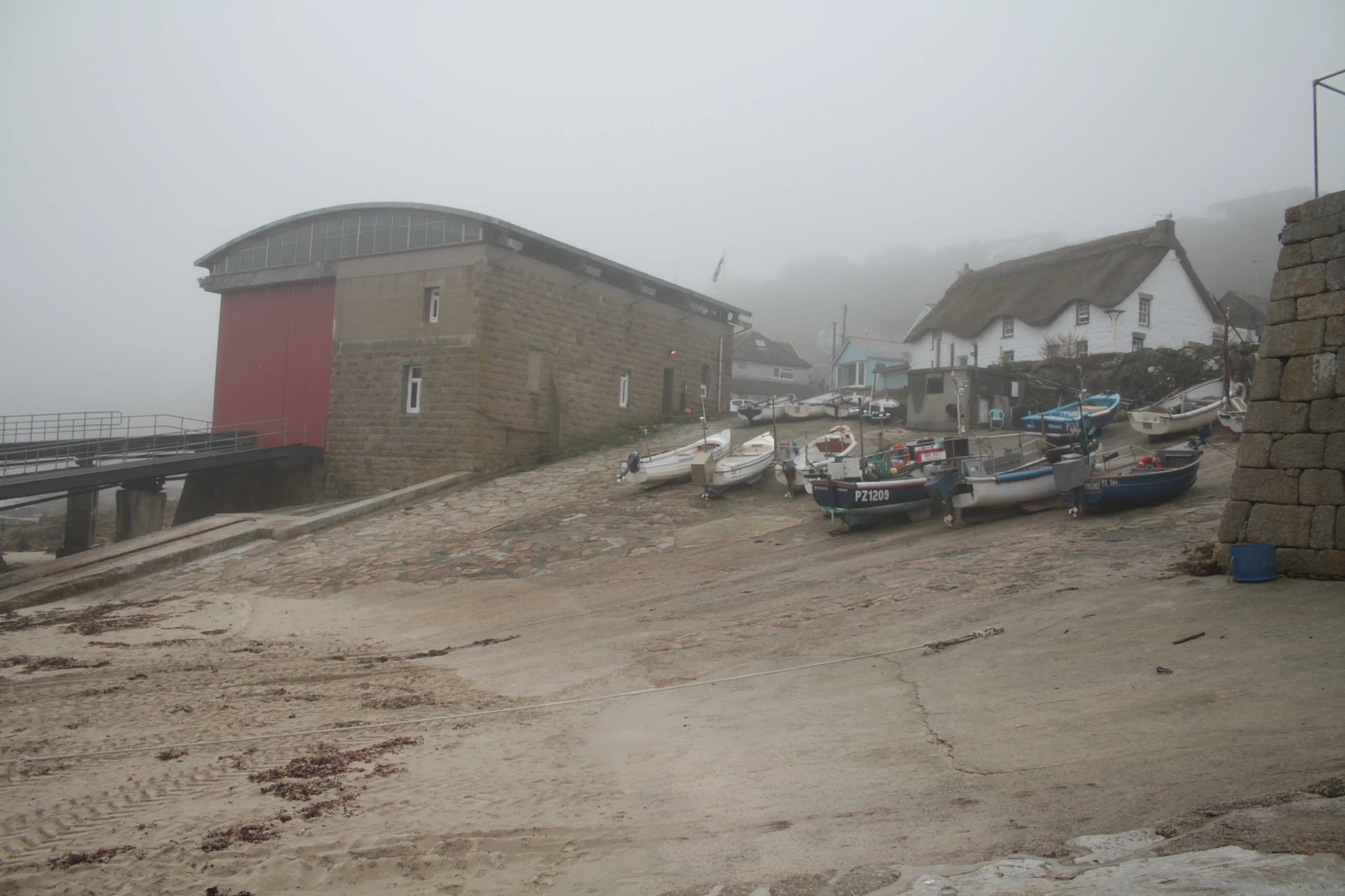 Foggy coastal scene with small fishing boats on a ramp near stone boathouse and thatched cottage