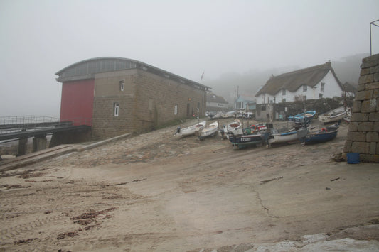 Foggy coastal scene with small fishing boats on a ramp near stone boathouse and thatched cottage