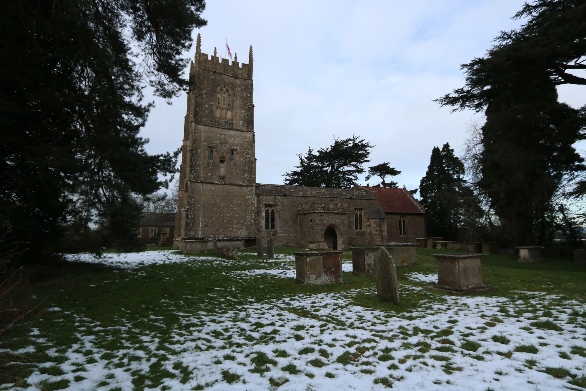 Historic Wickwar Church in winter with light snow on grass and surrounding gravestones, under a cloudy sky