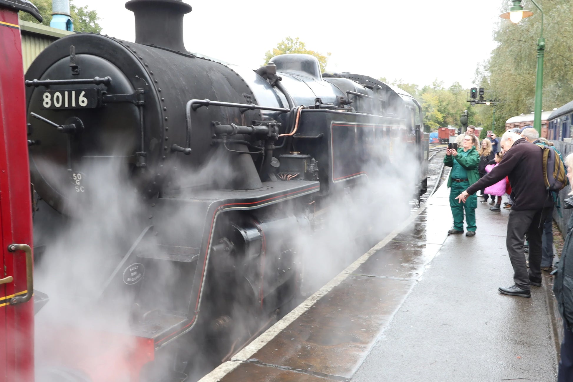 Black vintage steam locomotive 80116 releasing steam at a train station platform with people observing and taking photos