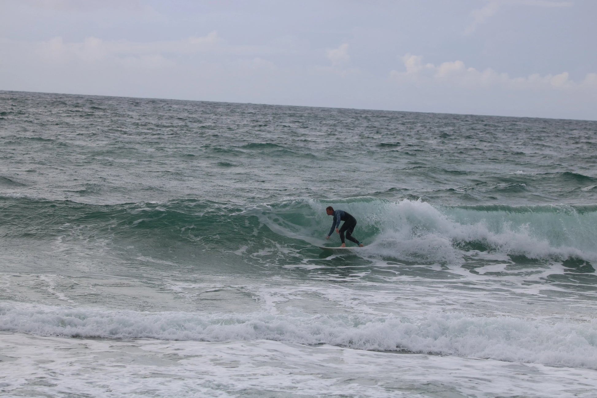 Surfer in a wetsuit riding a green wave at Praa Sands beach with cloudy sky