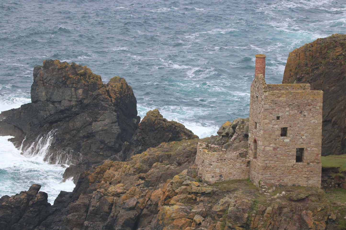 Historic stone building with chimney on rocky Cornwall coastline by rough sea waves
