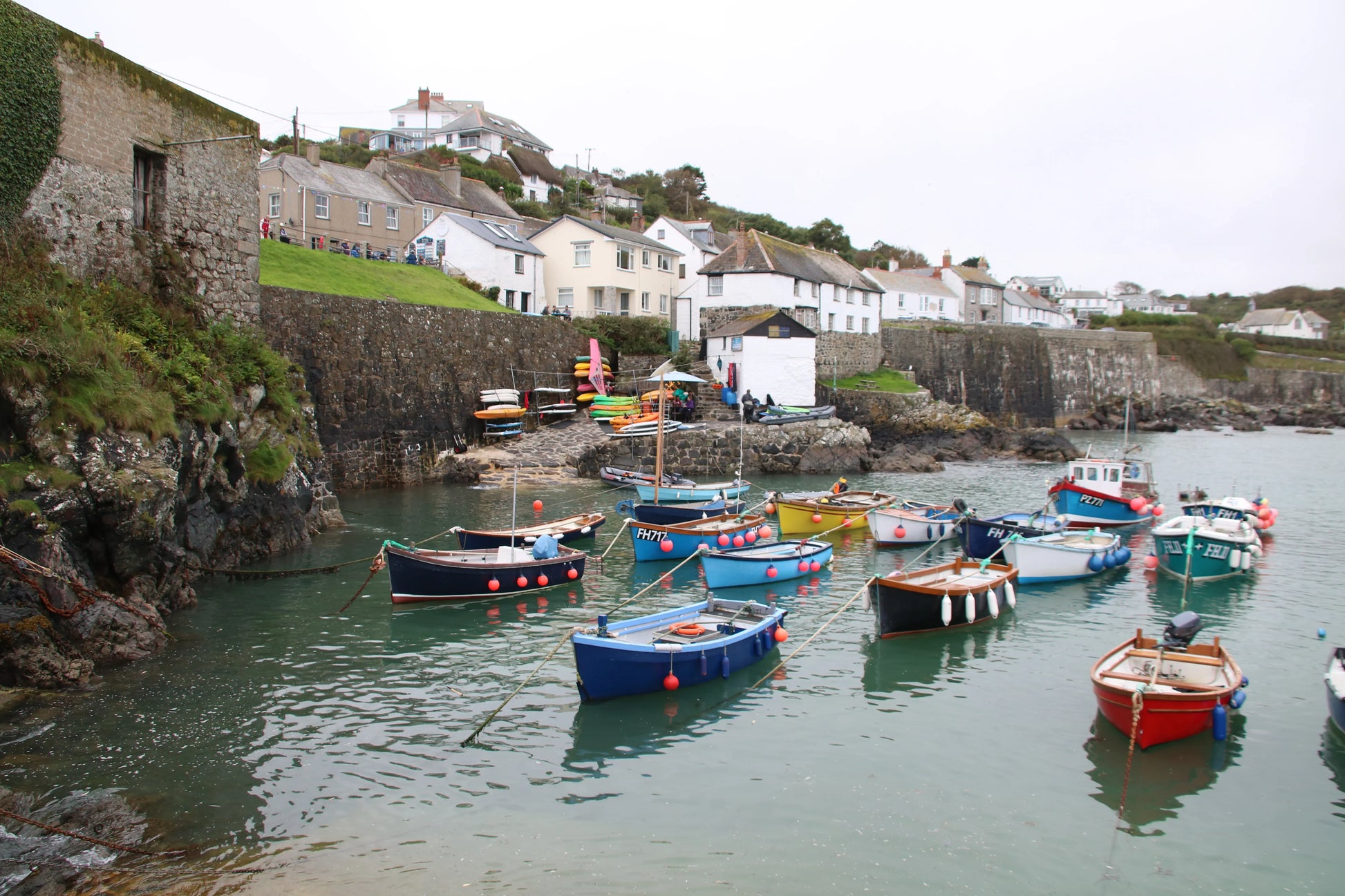 Colorful fishing boats moored in a small harbor with stone walls and coastal houses in the background