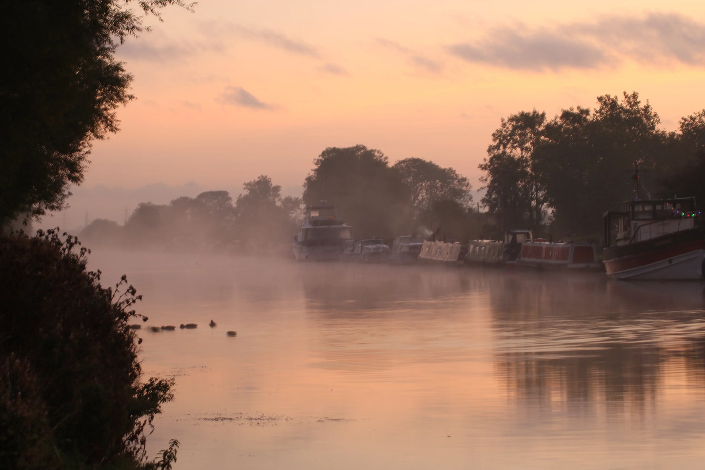 Mist rising over calm river at sunrise with moored boats and trees along the bank, soft pastel sky