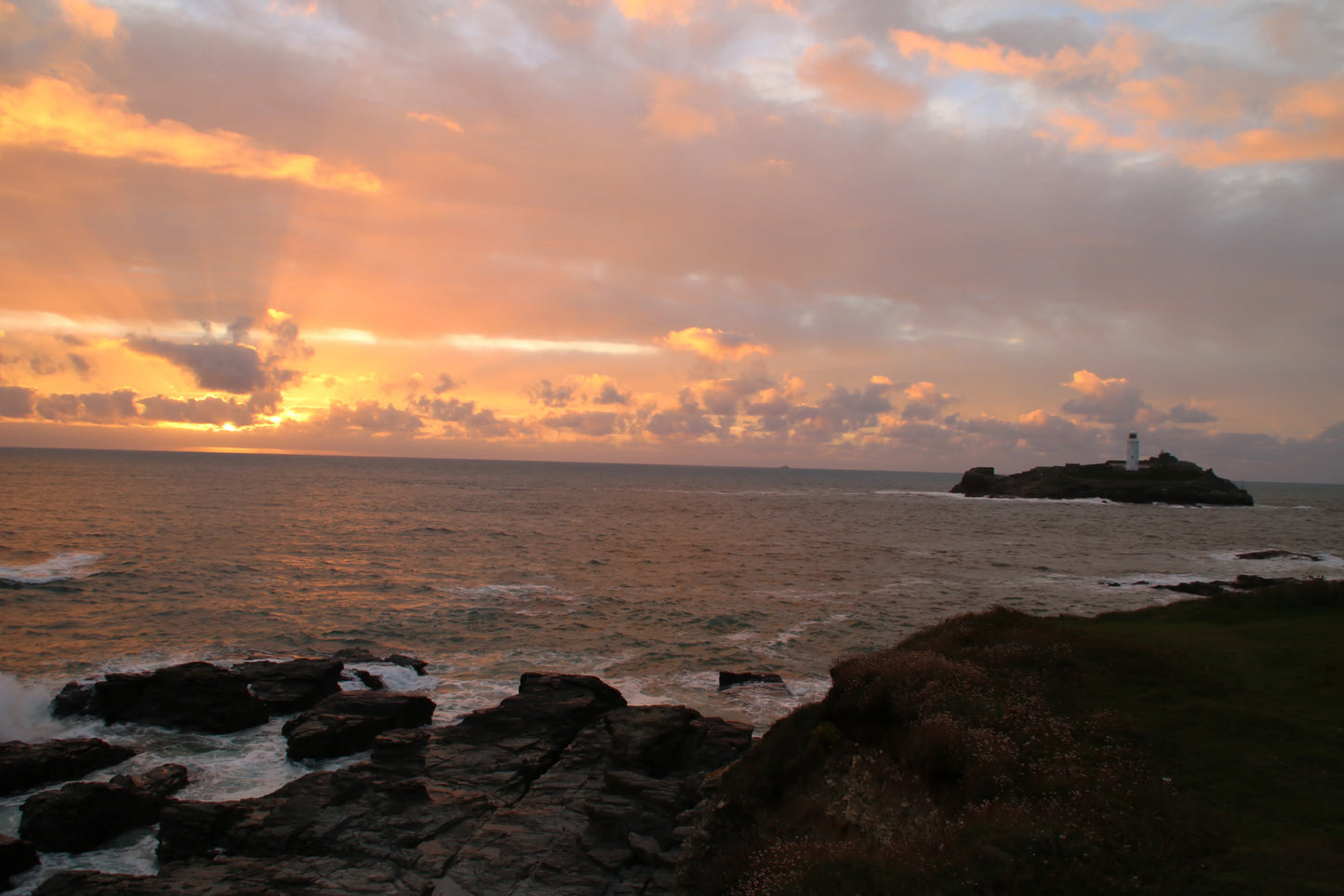 Sunset over rocky coastline with waves and a lighthouse on a small island