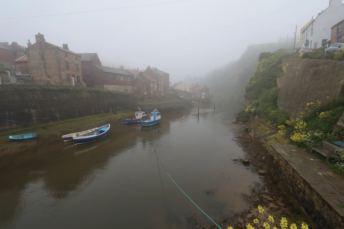 Foggy coastal village harbor with moored boats, stone buildings, and lush greenery in Yorkshire