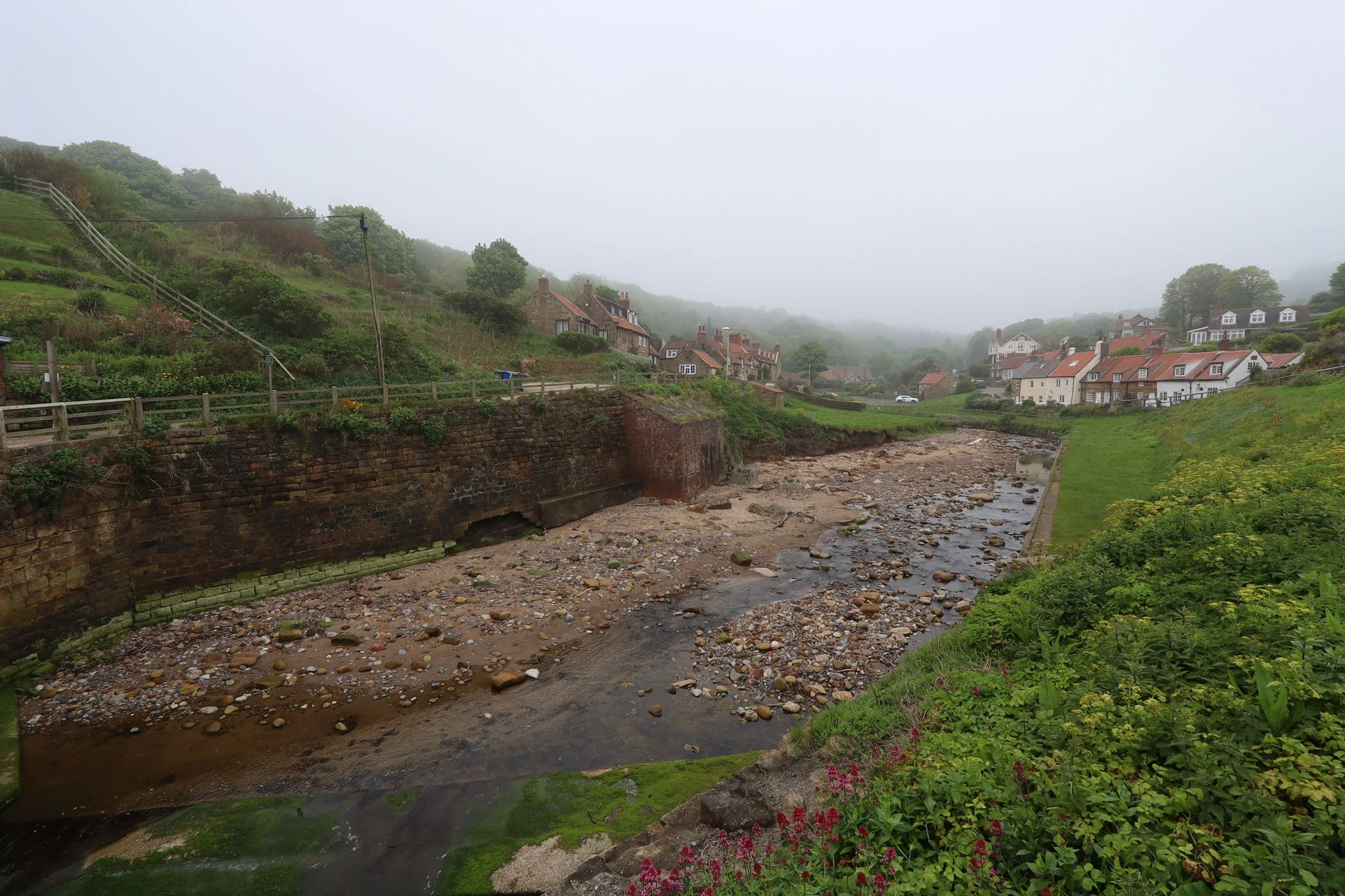 Foggy village landscape with stone cottages, a rocky stream, and lush green vegetation in British countryside