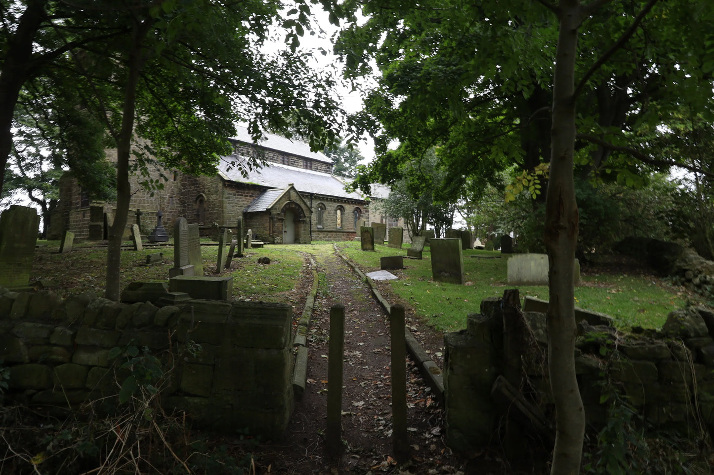 Stone church with slate roof surrounded by lush green trees and old gravestones in a shaded graveyard