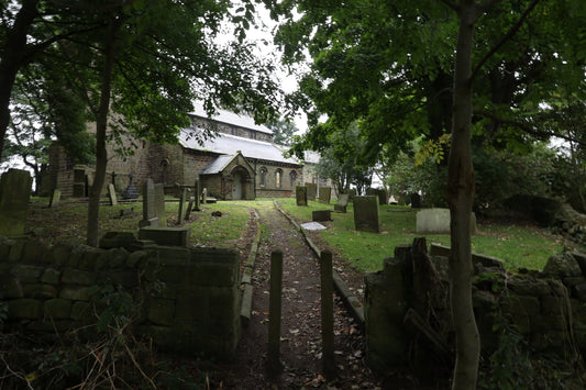 Stone church with slate roof surrounded by lush green trees and old gravestones in a shaded graveyard