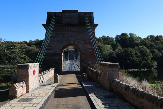 Historic stone and metal suspension bridge over river with trees under clear blue sky