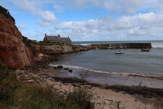 Coastal harbor with stone cottages on rocky cliffs, fishing boats docked in calm sea under cloudy sky