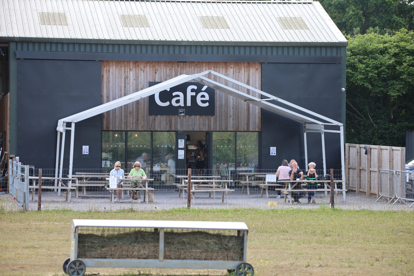Outdoor café seating with wooden picnic tables and customers in front of a modern barn-style building
