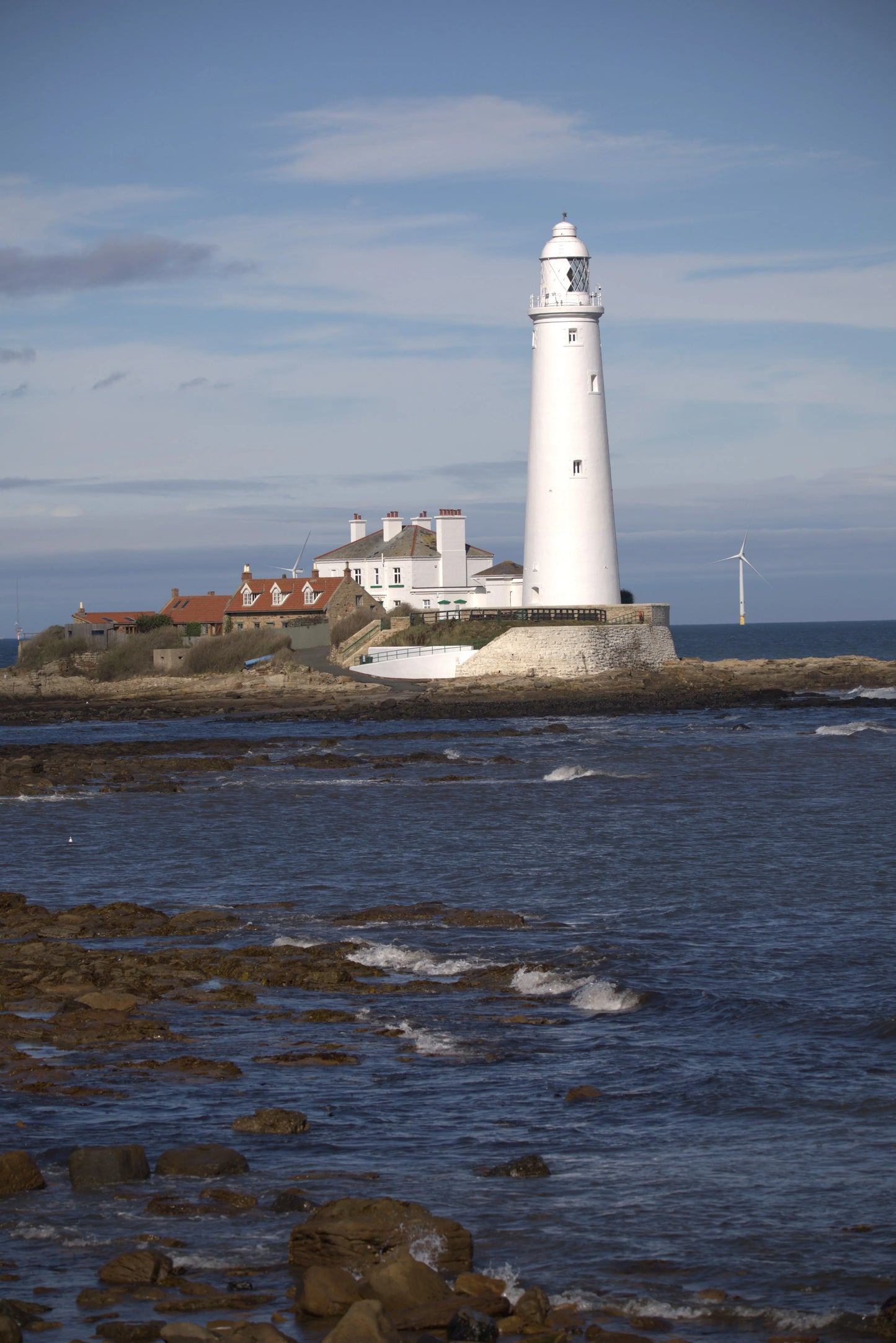St Mary's Lighthouse on rocky coast with sea waves and wind turbines under a partly cloudy sky