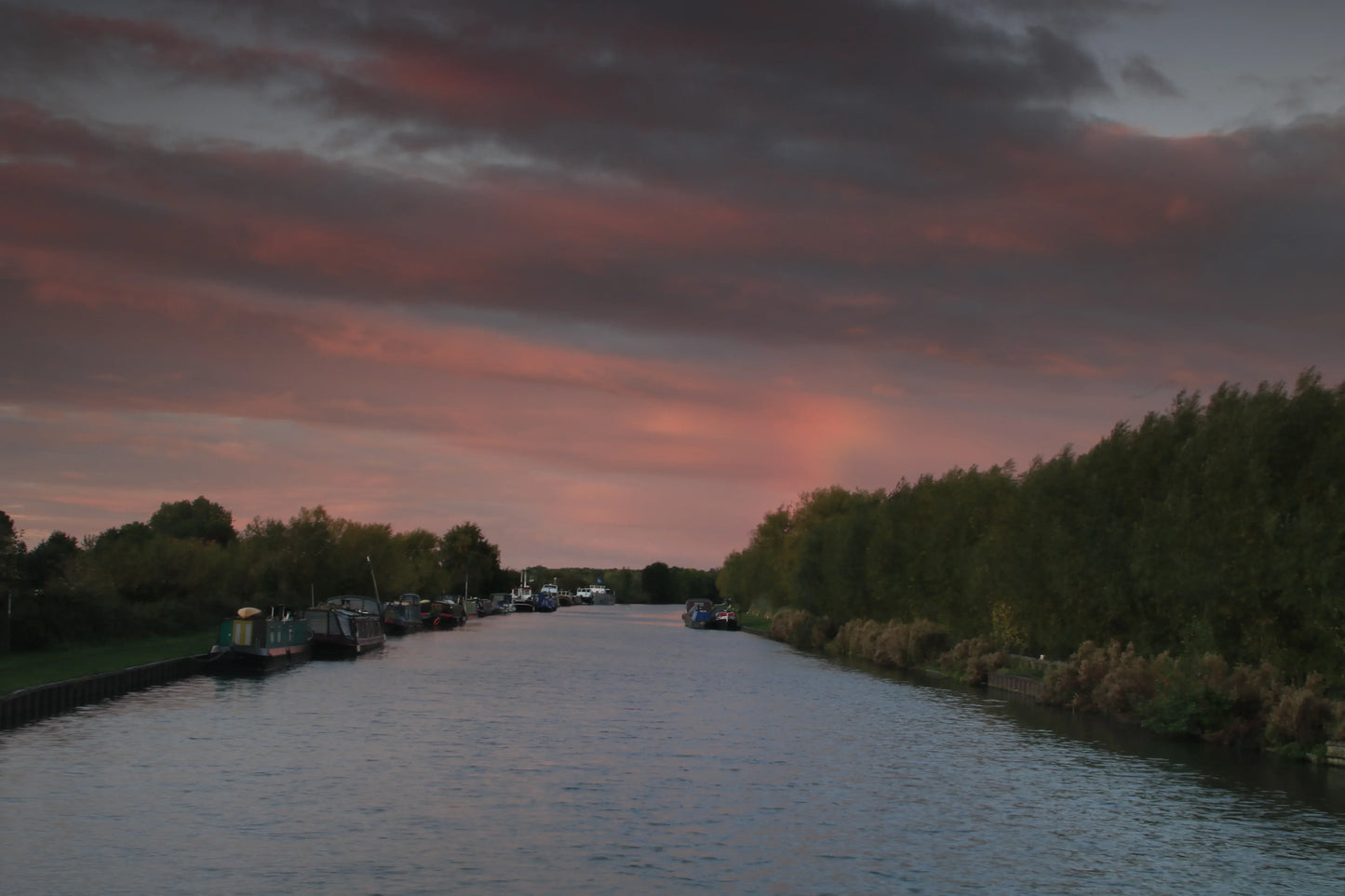 British canal with moored boats and dense trees at sunset under dramatic pink and gray sky