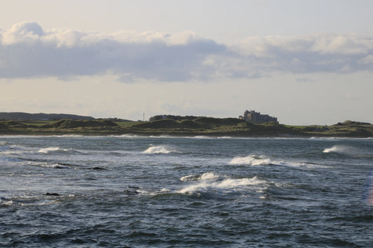 British coastline with choppy sea waves, grassy dunes, and historic castle in the distance under cloudy sky