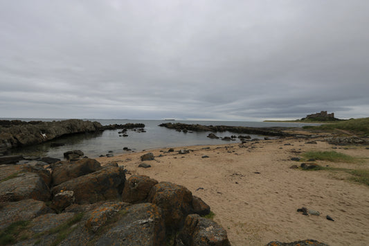 Coastal landscape with sandy beach, rugged rocks, calm sea, and distant castle under overcast sky