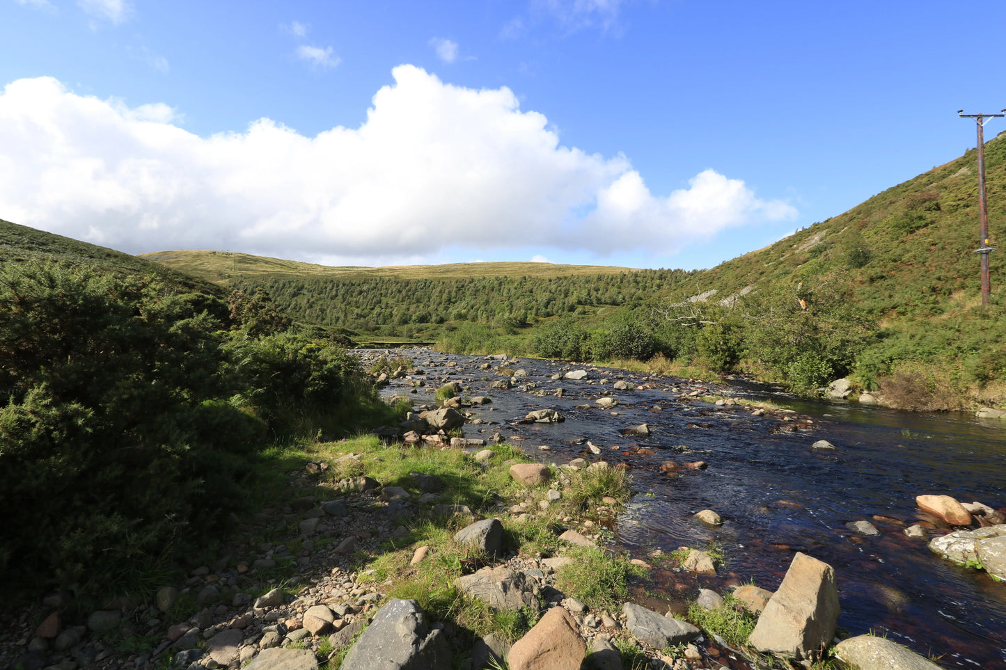 Rocky river flowing through green hills under a blue sky with white clouds