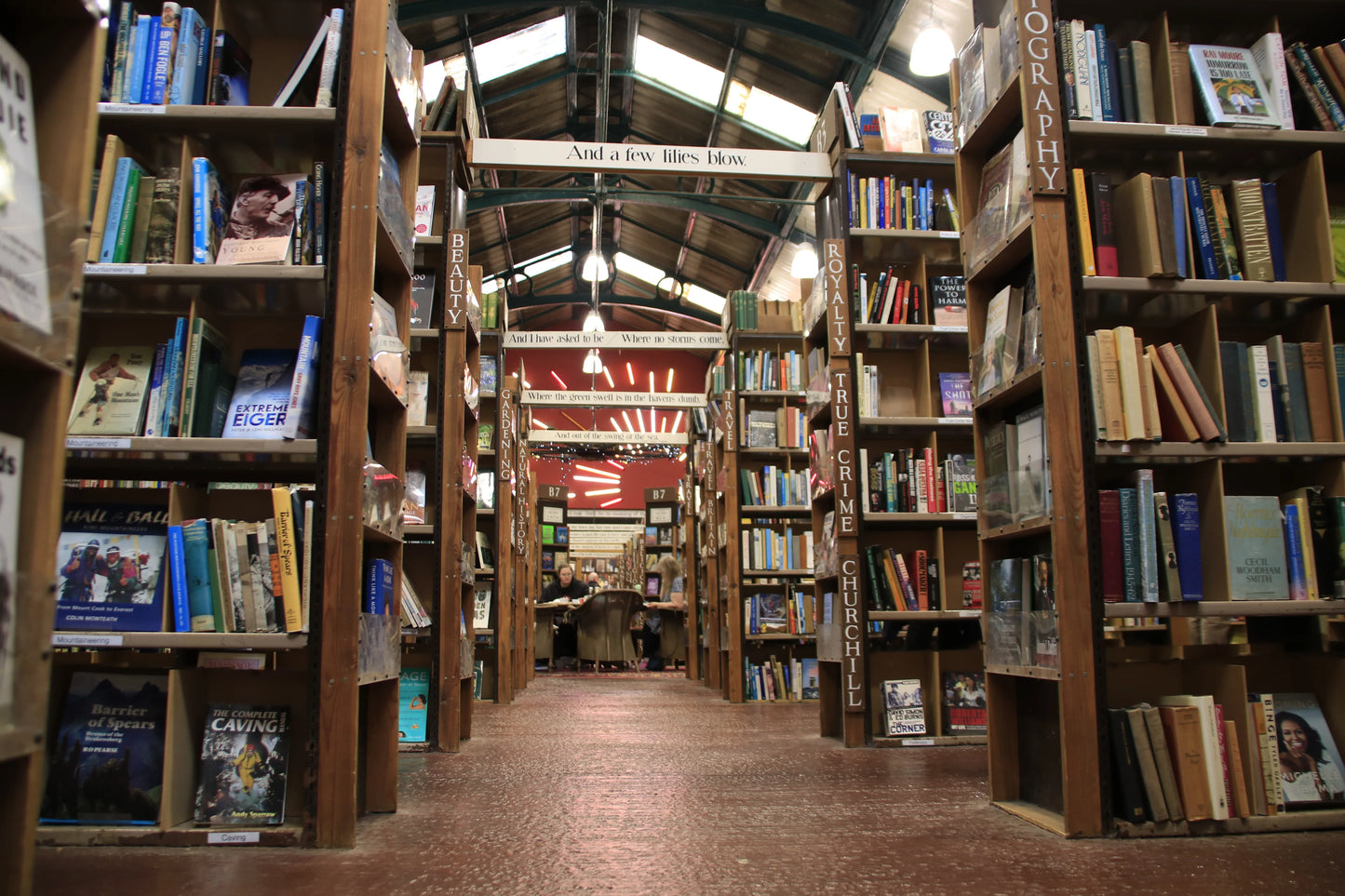 Interior aisle of a cozy bookstore with wooden shelves labeled by genres and two people sitting at a table in the background