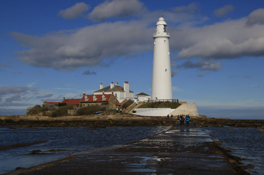 St Mary's Lighthouse on rocky Northumberland coast with visitors walking along wet causeway under blue sky