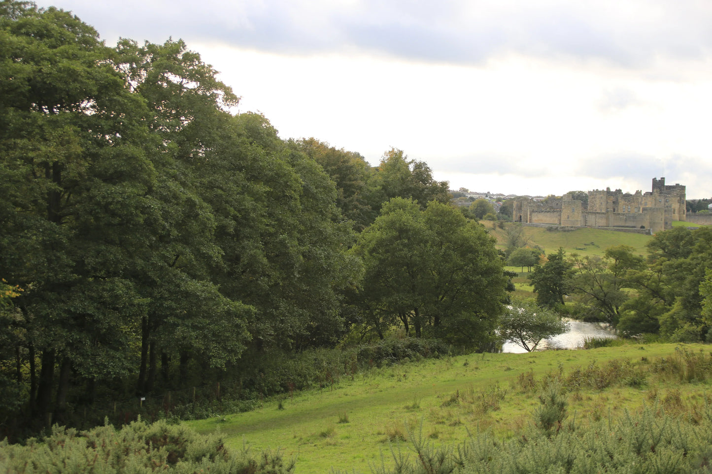 Alnwick Castle in Northumberland viewed from green countryside with trees and a small river under cloudy sky
