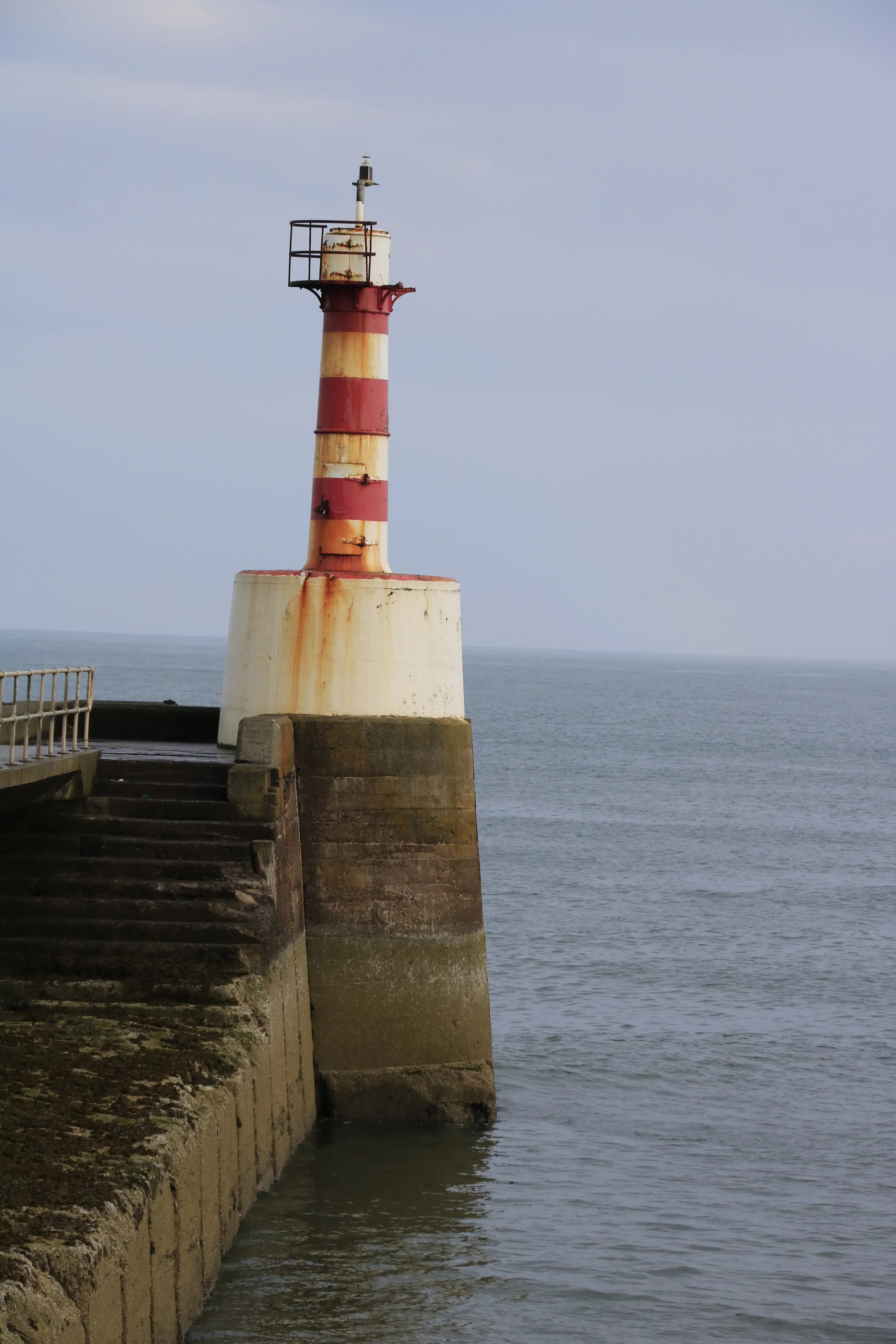 Red and white striped lighthouse on concrete pier by calm sea under overcast sky