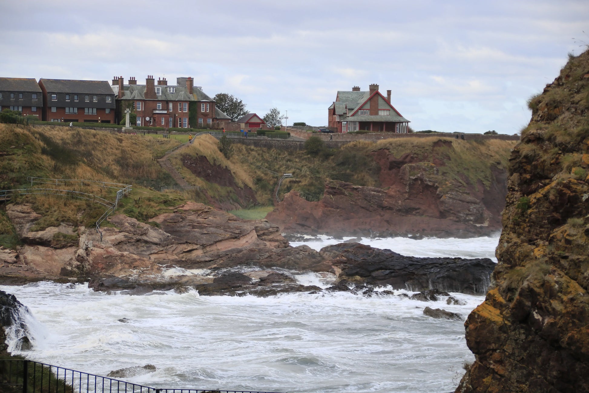 Northumberland coastal landscape with rocky cliffs, turbulent sea waves, and historic buildings on grassy cliff tops in Dunbar, UK
