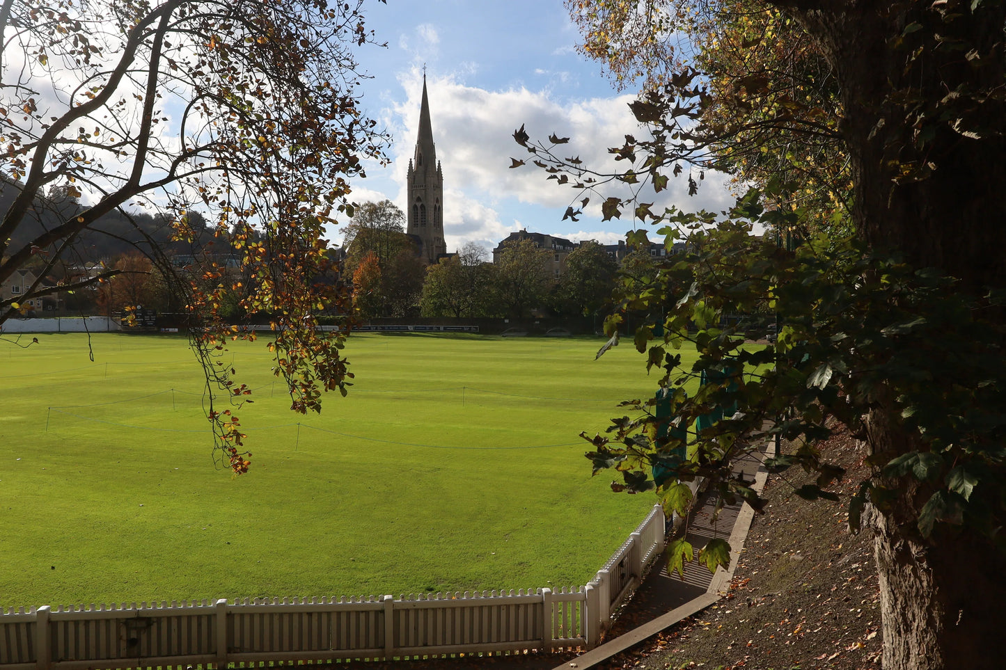 Green cricket field with white fence framed by autumn trees, Bath Cathedral spire in background