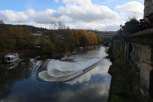 Autumn landscape of Bath weir with river, cascading water, boats, and historic stone balustrade under cloudy sky