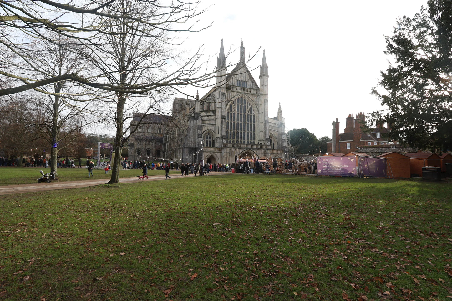 Winchester Cathedral with Christmas market stalls and visitors on green lawn under bare trees